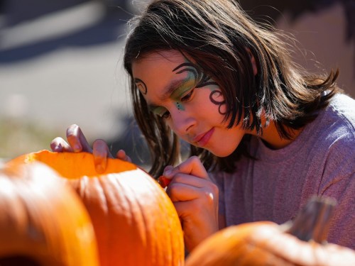 A young person with face paint carves a pumpkin, focused and surrounded by bright orange pumpkins.