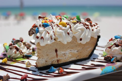 A slice of ice cream pie topped with whipped cream, chocolate drizzle, and colorful candy, set against a beach background.