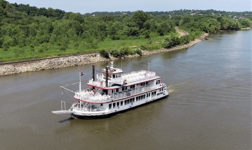 A riverboat cruises along a calm river, surrounded by lush greenery and a railway track in the background.