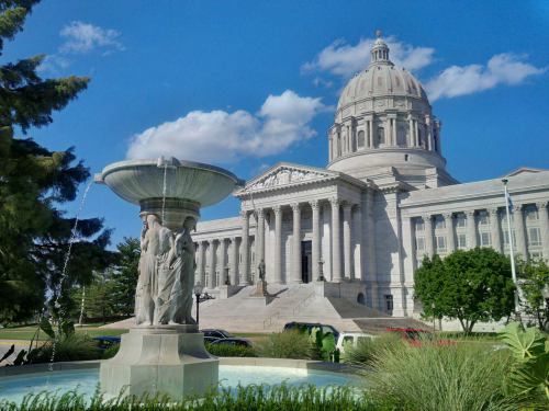 A grand stone building with a dome, featuring a fountain in the foreground and blue skies above.