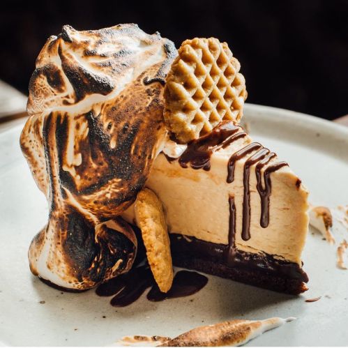 A slice of cheesecake topped with toasted meringue, chocolate drizzle, and a waffle cookie on a plate.