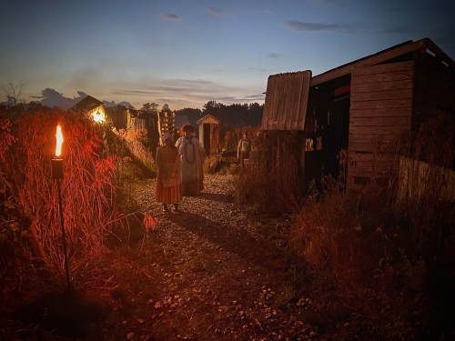 Two figures walk down a gravel path surrounded by rustic buildings and tall grass, illuminated by a torch at dusk.