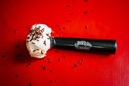 A black ice cream scoop with a scoop of vanilla ice cream topped with chocolate sprinkles on a red background.