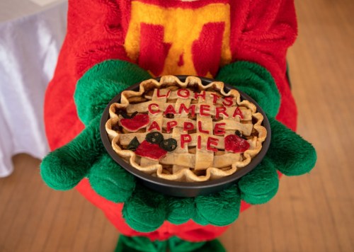 A person in a colorful costume holds a pie decorated with the words "Lights, Camera, Apple Pie" and fruit designs.