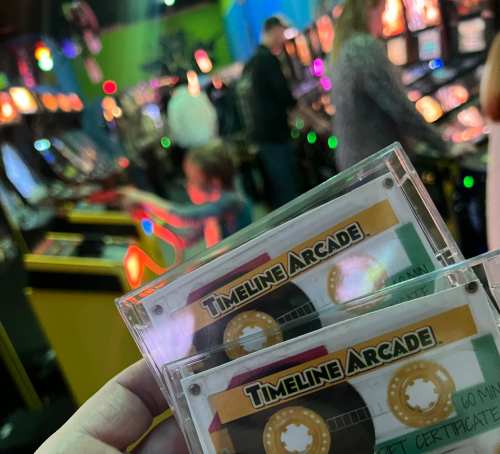 A hand holds two arcade tokens labeled "Timeline Arcade" in a colorful arcade filled with games in the background.