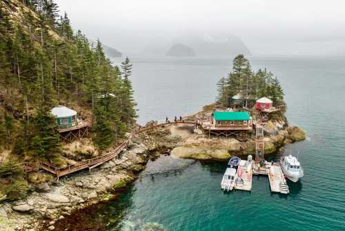 Aerial view of colorful cabins on a rocky shore, surrounded by trees and boats docked in calm waters. Foggy mountains in the background.