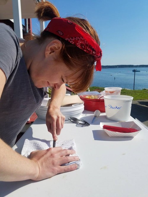 A woman in a red bandana works on a table by the water, focused on a task with tools and containers nearby.