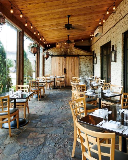 A cozy restaurant patio with wooden tables, chairs, and string lights, featuring a stone floor and rustic decor.