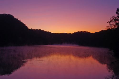 A serene lake at sunrise, reflecting vibrant purple and orange hues with mist rising from the water.