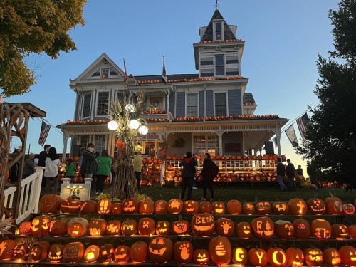 A decorated house with numerous carved pumpkins on the steps, surrounded by people enjoying a festive atmosphere.