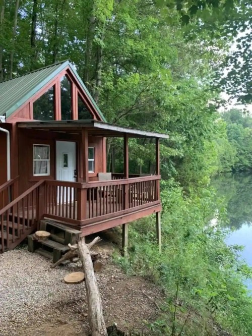 A cozy red cabin with a porch overlooking a serene river, surrounded by lush green trees.