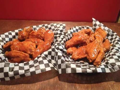 Two baskets of chicken wings with a glossy sauce, served on checkered paper. One basket has more wings than the other.