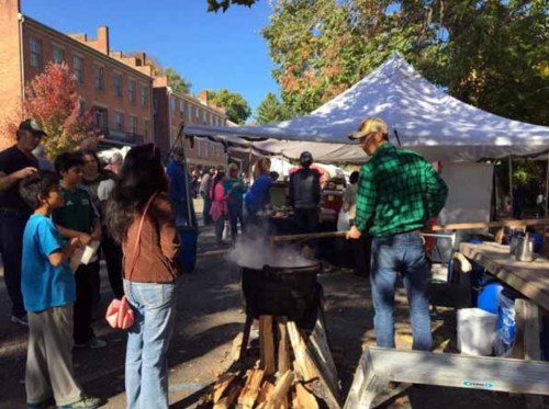A crowd gathers at an outdoor market, watching a person cooking over a fire in a large pot. Tents and trees are in the background.