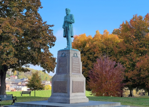 A bronze statue of a man in a coat stands on a pedestal, surrounded by colorful autumn trees.