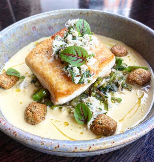 A beautifully plated dish featuring a crispy fish fillet atop greens, surrounded by a creamy sauce and garnished with herbs.