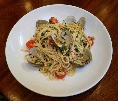 A plate of linguine pasta topped with clams, cherry tomatoes, herbs, and breadcrumbs on a wooden table.