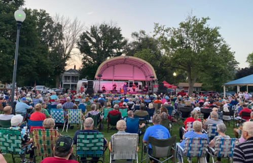 A large crowd sits in lawn chairs at an outdoor concert, with a stage and trees in the background during sunset.