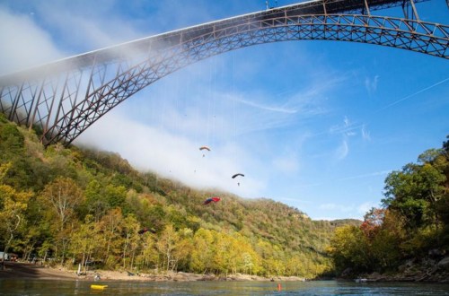 A scenic view of a bridge over a river, with colorful parachutes descending against a backdrop of autumn trees.