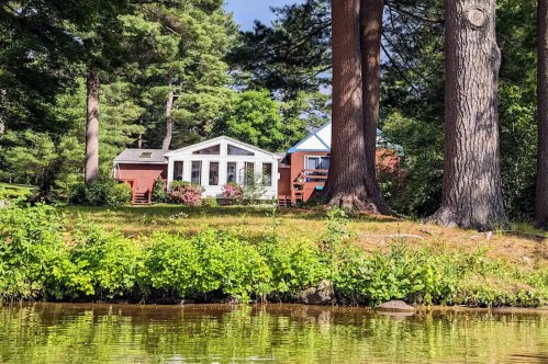 A serene lakeside view of a house surrounded by trees and greenery, reflecting a peaceful summer atmosphere.
