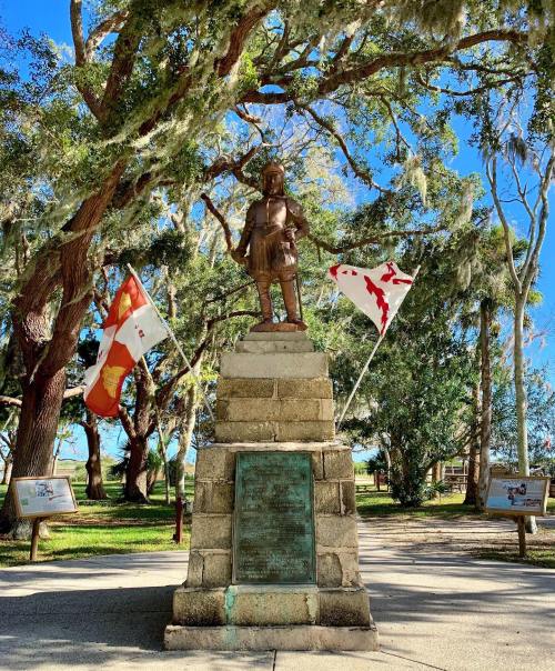 A bronze statue of a historical figure stands on a stone pedestal, surrounded by trees and flags in a park setting.