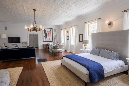 Spacious bedroom with a large bed, chandelier, and dining area, featuring bright windows and stylish decor.