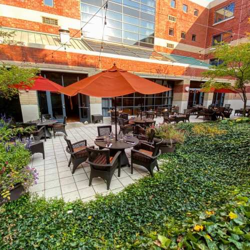 Outdoor dining area with tables under orange umbrellas, surrounded by greenery and flowers in a brick building setting.