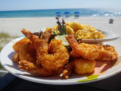A plate of fried shrimp with a side of macaroni salad, set against a beach backdrop with blue umbrellas.