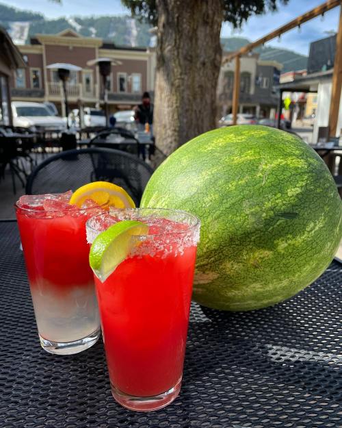 Two colorful cocktails with garnishes sit on a table next to a large watermelon in a sunny outdoor setting.