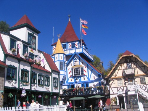 Colorful buildings with unique architecture in a quaint village square under a clear blue sky. Flags are flying above.
