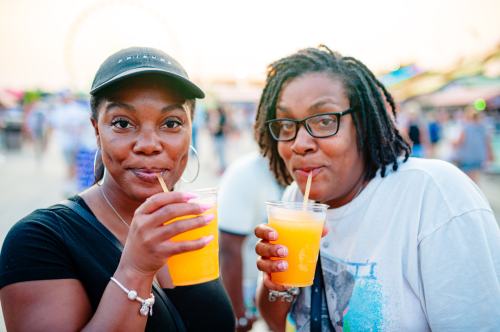 Two women smile while holding orange drinks with straws at a fair, with a ferris wheel in the background.