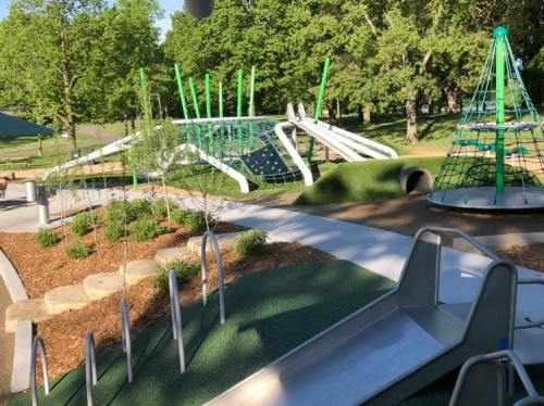 A modern playground featuring slides, climbing structures, and green landscaping surrounded by trees.