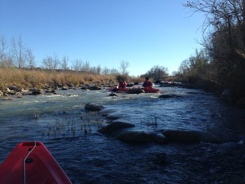Two kayakers navigate a rocky river under a clear blue sky, surrounded by sparse vegetation and trees.