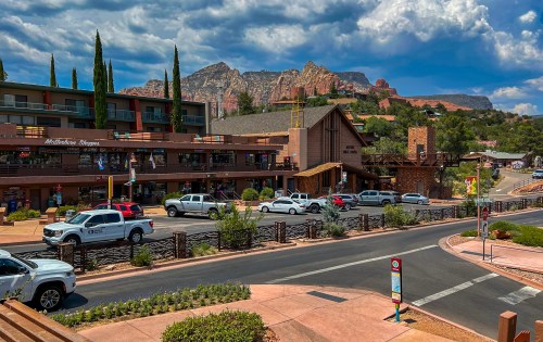 A scenic view of a shopping area with mountains in the background, featuring cars and greenery under a cloudy sky.