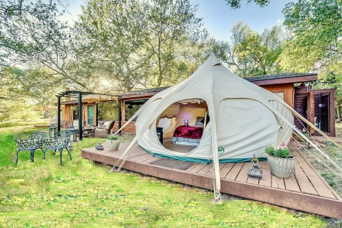 A cozy tent on a wooden deck surrounded by greenery, with nearby seating and a rustic cabin in the background.