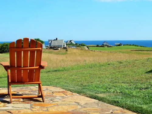 A wooden chair overlooks a grassy field and ocean, with houses in the distance under a clear blue sky.