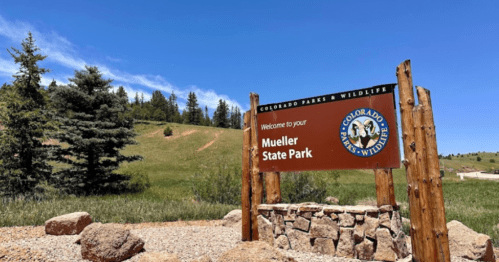 Welcome sign for Mueller State Park, surrounded by trees and grassy hills under a clear blue sky.