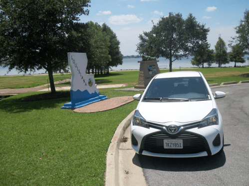A white car parked near a Louisiana state sign by a lake, with trees and a blue sky in the background.
