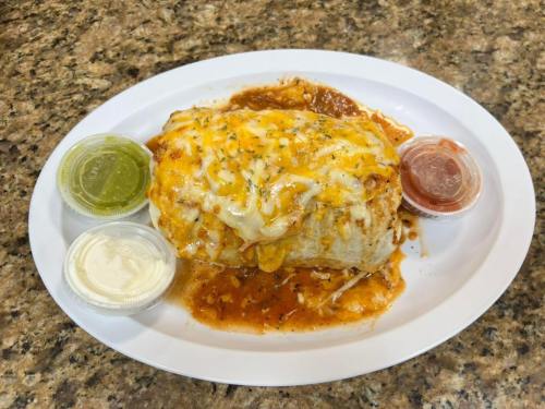 A cheesy burrito smothered in sauce, served on a plate with three dipping sauces: green, sour cream, and red.