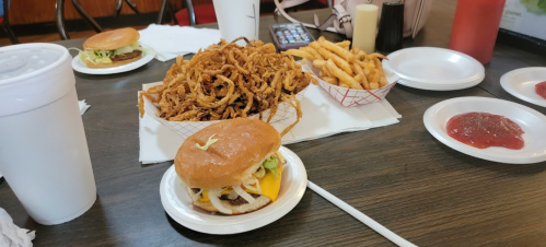 A table with two burgers, a large pile of onion rings, fries, and condiments in a casual dining setting.