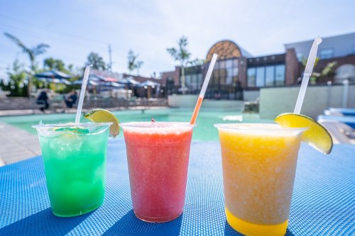 Three colorful frozen drinks in clear cups sit on a table by a pool, with a sunny sky and lounge chairs in the background.