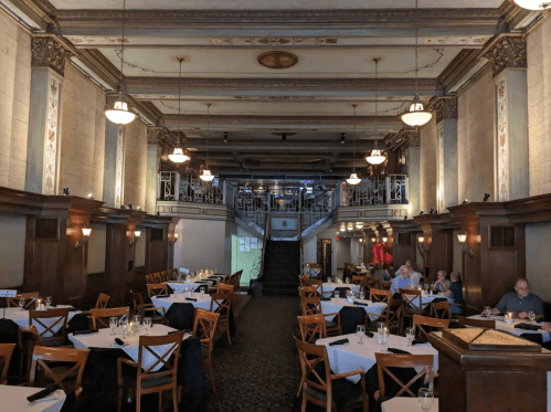 Elegant restaurant interior with wooden furniture, high ceilings, and a staircase leading to an upper level.