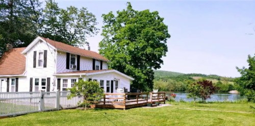 A charming white house with a deck, surrounded by greenery, near a calm lake and rolling hills in the background.