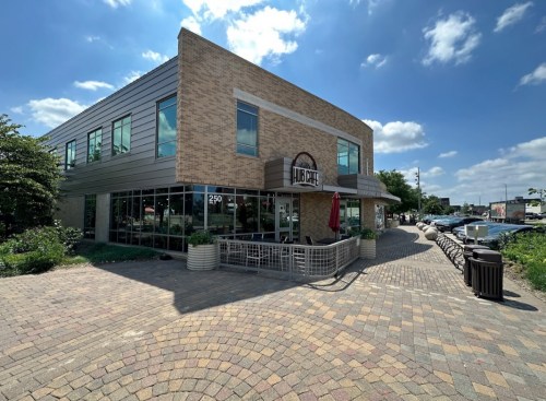 A modern café building with large windows, outdoor seating, and a brick pathway under a blue sky with clouds.