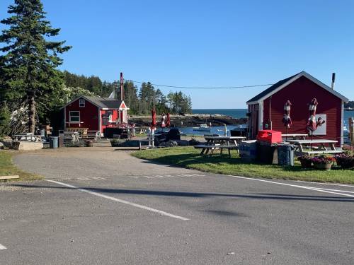 A scenic view of a coastal area with red cabins, picnic tables, and trees under a clear blue sky.