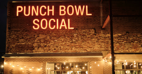 Neon sign reading "PUNCH BOWL SOCIAL" on a brick building, illuminated at night with string lights in the foreground.