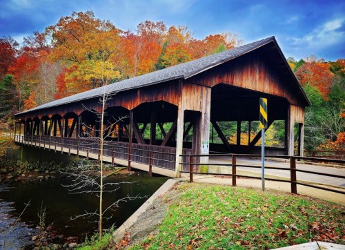 A wooden covered bridge spans a river, surrounded by vibrant autumn foliage and a clear blue sky.