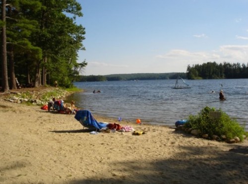 A sandy beach by a lake with people swimming, lounging on chairs, and trees in the background under a clear sky.