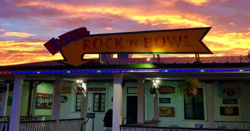 A colorful sunset behind the "Rock 'n Bowl" entrance sign, with vibrant lights illuminating the building.