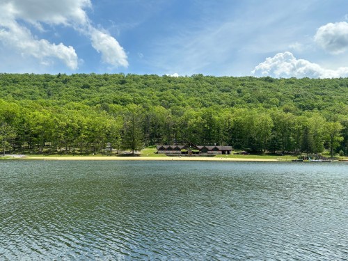 A serene lake surrounded by lush green trees and a distant cabin under a blue sky with fluffy clouds.