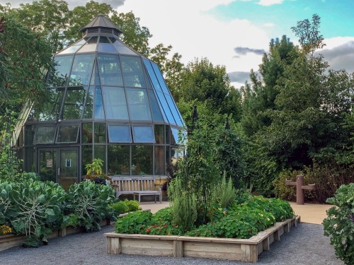 A glass greenhouse surrounded by lush greenery and garden beds, under a partly cloudy sky.